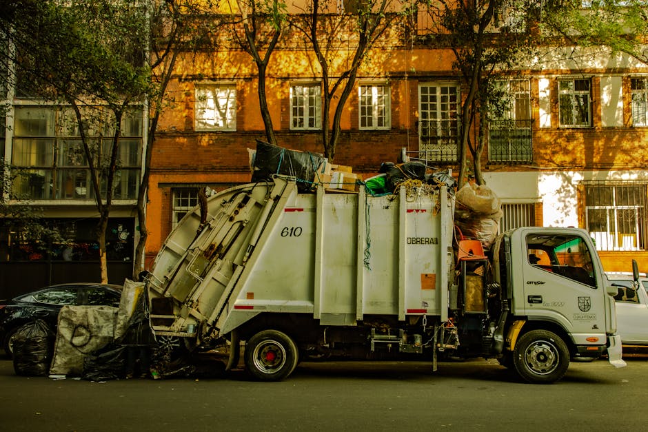 A pile of mixed waste materials is shown on the ground outdoors, consisting of black plastic trash bags, a yellow plastic container, large discarded mattress parts with a beige fabric cover, and vario