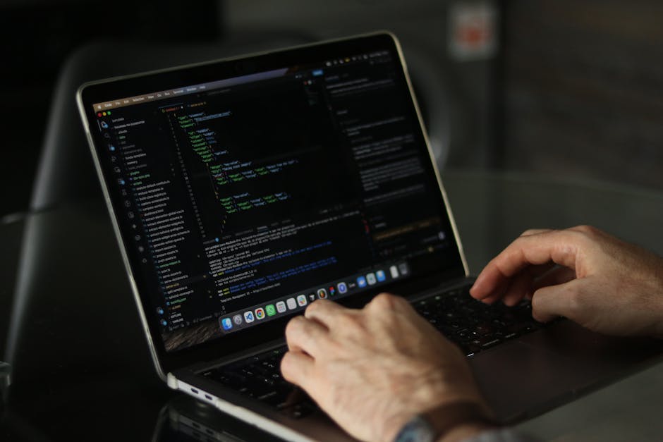 The image depicts a person seated at a desk, viewed from behind, working on a laptop with a dark screen displaying multicolored lines of code. The individual, wearing a light blue shirt, is using a mo